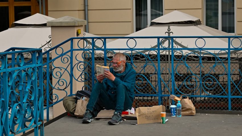 Homeless man reading a book on a city street in Kraków, sitting by a blue railing with a cardboard sign asking for help.