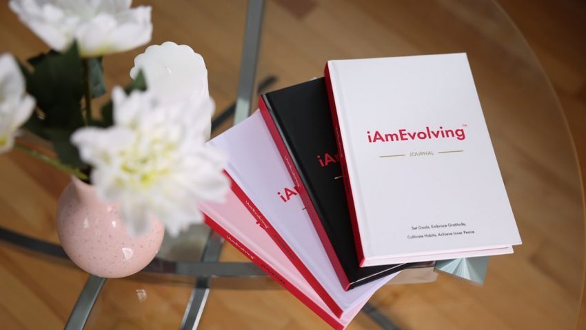 A stack of minimal hardcover journals arranged on a glass table in a bright indoor setting. Soft natural light highlights clean lines, neutral tones, and simple design details, while a small floral accent adds balance and texture to the composition. The image conveys calm, organization, and intentional living, making it suitable for concepts related to journaling, personal growth, reflection, stationery, lifestyle design, and mindful everyday environments.